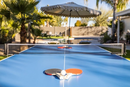 A blue outdoor table tennis table with two paddles and a ball resting in the center. An umbrella and lounge area are visible in the background.