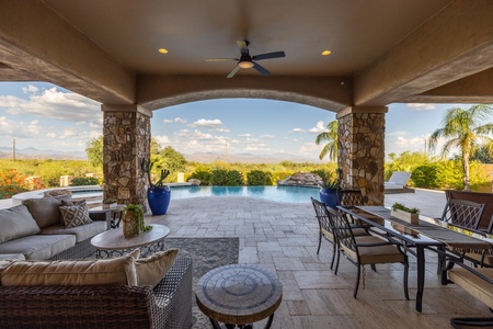 Covered outdoor patio with seating area and table, overlooking a pool and distant mountains.