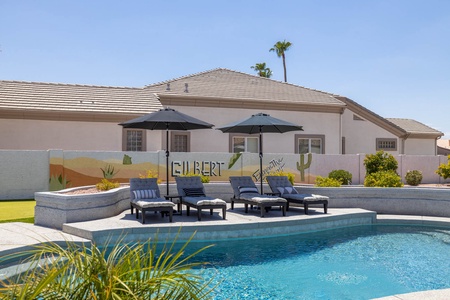 A backyard with a swimming pool, lounge chairs under umbrellas, and a mural on the wall depicting a desert scene with the word "Gilbert" and cacti.