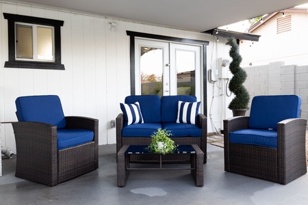 Outdoor patio setup with two armchairs and a loveseat, all with blue cushions, a glass-top coffee table with a plant, set against a white-painted house with a glass French door.