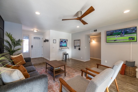 A modern living room with a ceiling fan, a wall-mounted TV displaying a baseball game, a gray sofa, two gray chairs, a wooden coffee table, and a plant in the corner. The room has wooden flooring.