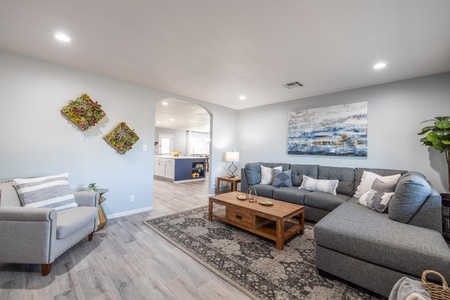 A modern living room with a gray sectional sofa, wooden coffee table, and armchair. Wall art and plant decor are visible. An archway leads to a kitchen in the background.