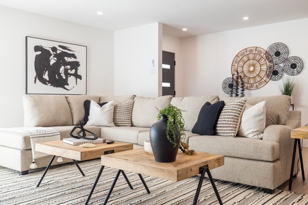 A modern living room with a beige sectional sofa, wooden coffee tables, black and white decor, a rug with geometric patterns, wall art, and decorative woven baskets on the wall.