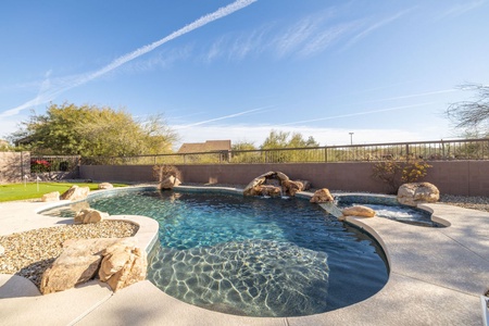 A backyard swimming pool with clear blue water, surrounded by a concrete deck and bordered by rocks and pebbles. Trees and a brick wall are visible in the background under a clear sky.