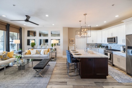 Modern open-concept kitchen and living room with white cabinetry, stainless steel appliances, a central island with barstools, and a comfortable seating area with grey and yellow accents.