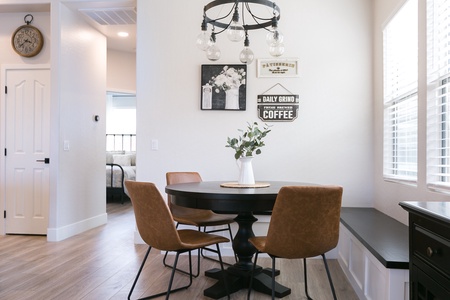 A small dining area with a round black table, four brown chairs, a vase with green leaves, and framed wall art. A black chandelier with bulb lights hangs above the table. Light streams in from windows.