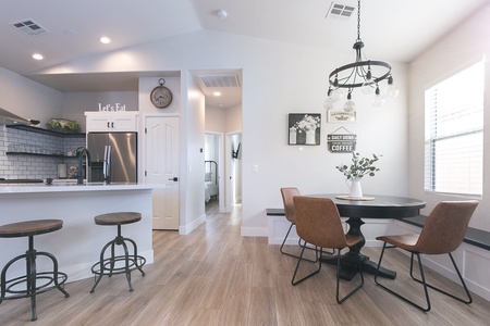 A modern kitchen and dining area with a white island, brown leather stools, a round dining table with brown chairs, a chandelier, and wall decor, next to a hallway leading to another room.