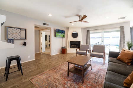 A living room with a couch, two chairs, a coffee table, and a wall-mounted TV. There is a fireplace with a round mirror above it, large windows with curtains, and a door leading outside.