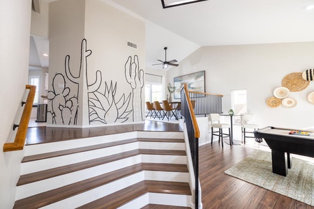 Modern, open-plan living space with wooden floors and staircase. Wall with cactus mural, dining area with six chairs, ceiling fan, and pool table on rug. Woven wall art and large painting decorate walls.