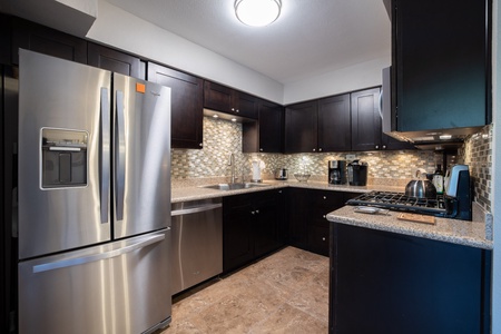 A modern kitchen features stainless steel appliances, dark wooden cabinets, granite countertops, a tiled backsplash, and bright overhead lighting.