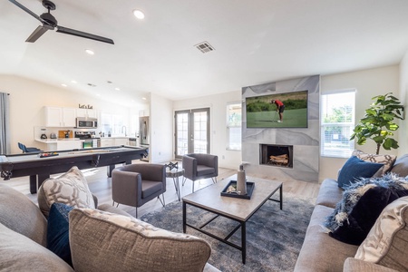 A modern living room with a sectional sofa, two armchairs, a coffee table, a wall-mounted TV, and a fireplace. The space includes a pool table, a ceiling fan, and a plant near the window.