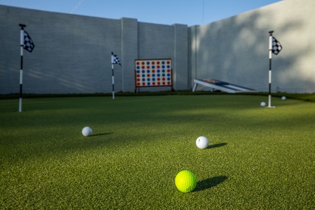 A small golf practice area with a green carpet, four checkered flag poles, several white golf balls, a yellow golf ball, a bean bag toss game board, and a Connect 4 game board nearby.