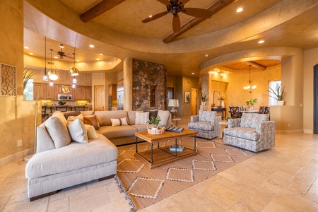 Spacious open-concept living room with a beige sectional sofa, patterned armchairs, a wooden coffee table, and a kitchen with wooden cabinetry and stone accents in the background.