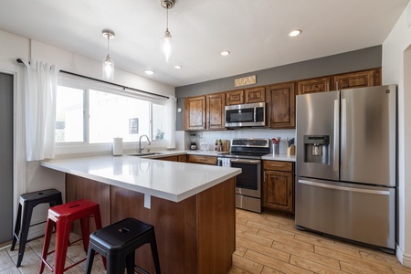 Modern kitchen with stainless steel appliances, wooden cabinets, a large window, and a white countertop island with three barstools.