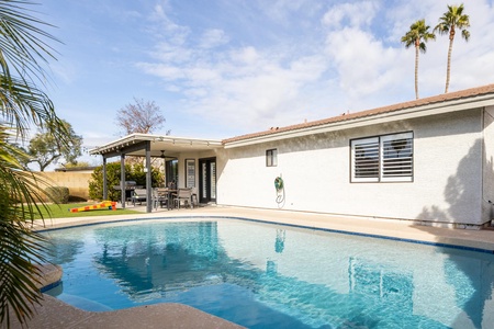 A house with a wooden patio, outdoor dining set, and a swimming pool in the backyard, with palm trees and a clear blue sky in the background.