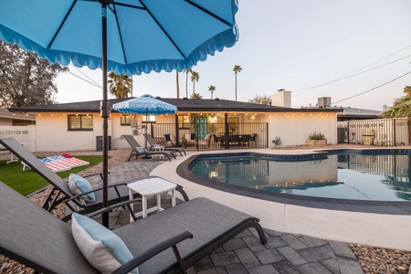 Backyard pool area with lounge chairs, tables under blue umbrellas, string lights, fenced pool, and a single-story house in the background.