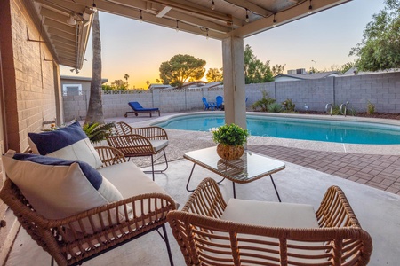 Outdoor patio with wicker seating, cushions, a glass-top table, and a potted plant overlooking a swimming pool with loungers, surrounded by a brick wall, during sunset.