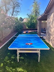 A blue ping pong table is set up outdoors on artificial grass, with two paddles placed in the center. Bags for cornhole are visible in the background on a sunny day.