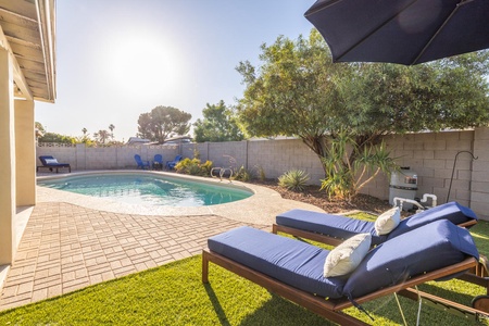 A backyard features a swimming pool, patio area, and two lounge chairs with blue cushions under an umbrella, surrounded by a brick wall and trees.