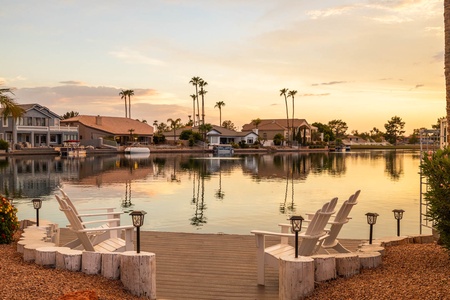 Lakeside view with four white Adirondack chairs on a wooden dock, surrounded by houses and palm trees, at sunset.