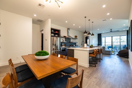 Modern open-concept kitchen and dining area with a wood table, blue chairs, a kitchen island with pendant lights, stainless steel appliances, and a living room with large windows in the background.