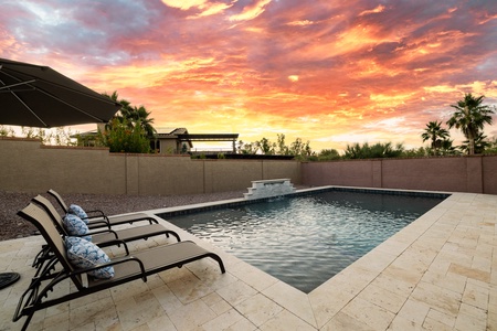 Four lounge chairs are positioned beside a rectangular pool under an umbrella, set against a backdrop of a sunset sky, with palm trees and a modern house enclosed by a stone wall.