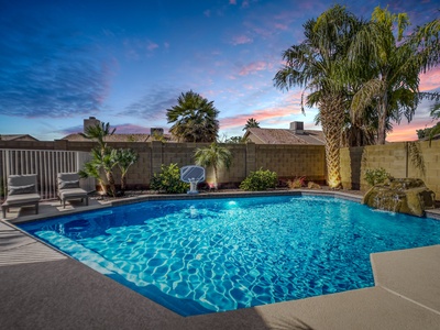 A backyard swimming pool with clear blue water is surrounded by lounge chairs, palm trees, and a stone wall. The sky is colorful at sunset.