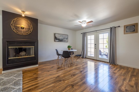 A modern living room features wooden flooring, a black fireplace, a round dining table with four chairs, and a large window with gray curtains. A ceiling fan and wall art are also visible.