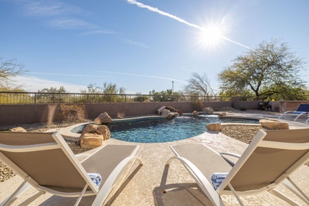 Two lounge chairs face a clear outdoor pool surrounded by rocks and desert vegetation under a bright, sunny sky.