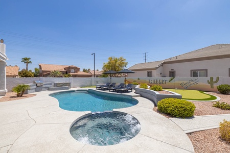 Backyard with a pool, hot tub, lounge chairs, and a putting green. Surrounding houses and clear blue sky are visible in the background.