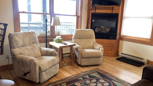 Two armchairs flank a wooden side table with a lamp and tissue box in front of a fireplace. A TV is mounted above the fireplace. Large windows let in light from outside. A patterned rug is on the floor.