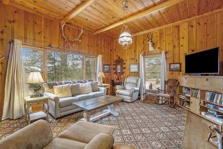 A cozy wooden living room with large windows, sofas, armchairs, a coffee table, a TV, and a bookshelf. The room is decorated with a hanging chandelier and animal-themed decor.