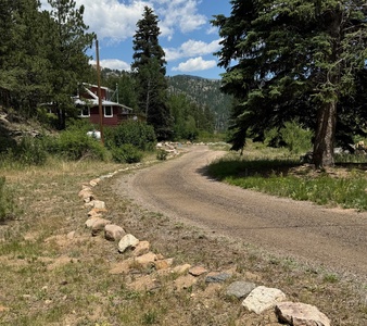 A gravel road curves past a wooden house surrounded by pine trees and greenery, with mountains visible in the background under a partly cloudy sky.