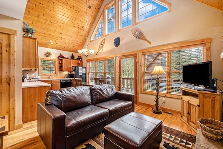 Living room with vaulted ceiling, leather sofa that seats 4, ottoman, floor lamp, flat screen tv, windows with views of outside, looking toward kitchen area.