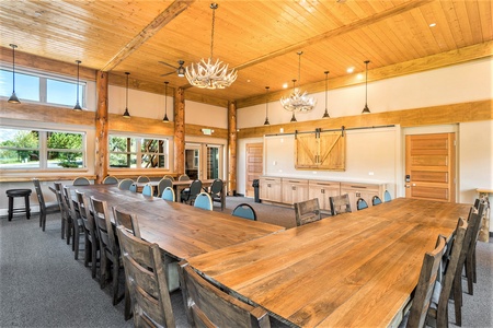 A rustic meeting room with long wooden tables, cushioned chairs, wooden walls, large windows, antler chandeliers, and a projector screen.