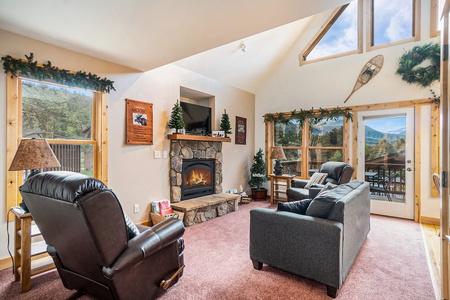 Living room with vaulted ceiling, sofa that seats 3, 2 armchairs, 2 side tables with lamps, fireplace, flat screen TV, and lots of windows with mountain views.