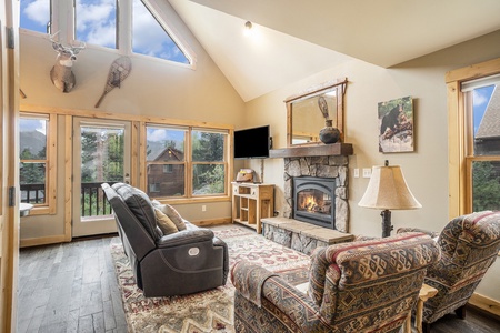 Living room with reclining sofa and two chairs, end table with lamp, flat screen TV, fireplace, and lots of windows with mountain views. Patio door leading to large deck.