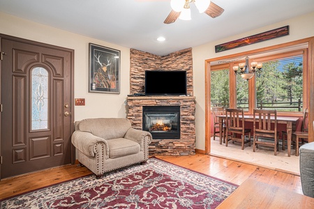 Living room with a stone fireplace, TV, armchair, and wooden dining table near large windows. A painting and a ceiling fan are also present.