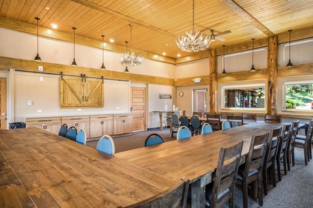 A spacious meeting room with wooden ceiling and floors, rectangular tables, blue chairs, and antler chandeliers. Large windows on the right offer a view of greenery outside.