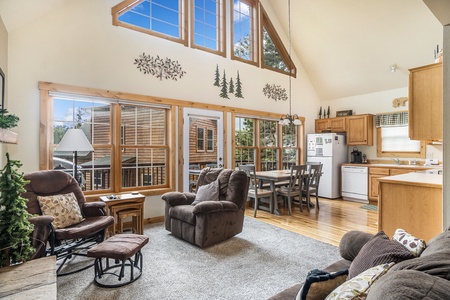 Living room with vaulted ceiling, sofa that seats 3, arm chair, chair with foot stool, looking into kitchen, and windows with mountain views.