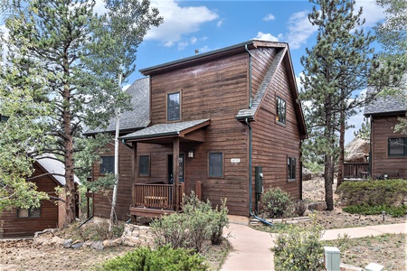 A two-story wooden cabin with a small front porch, surrounded by trees and a walkway in a natural setting.