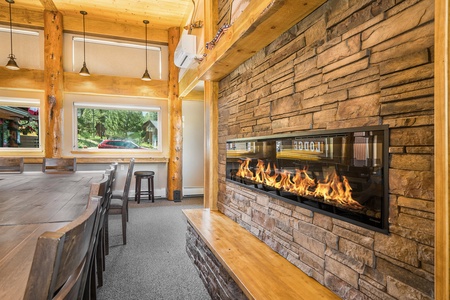 Rustic interior with a stone fireplace, wooden table, chairs, and large windows letting in natural light.