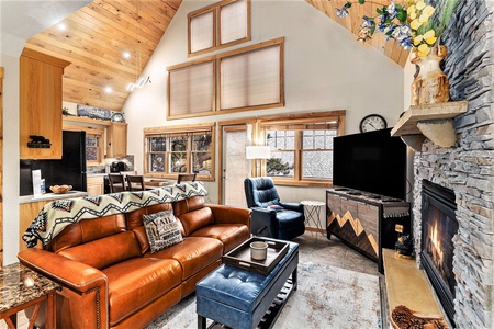 Cozy living room with a leather sofa, armchair, stone fireplace, and large TV. Wooden ceiling and walls add a rustic touch. Brightly lit with natural light from high windows.