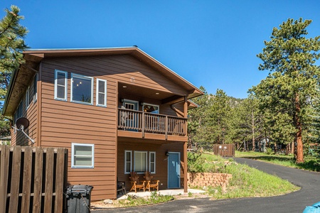 A two-story brown wooden house with a balcony, surrounded by trees. A paved driveway leads up to the house, and patio chairs are placed outside the lower-level entrance.