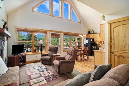 Open concept living room, with 2 reclining chairs, end table, flat screen TV, and lots of windows with view of mountains.