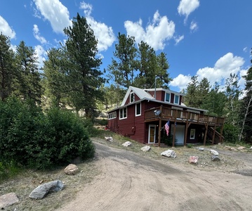 A red cabin with a wooden deck is surrounded by trees under a blue sky with clouds. An American flag is displayed at the entrance.