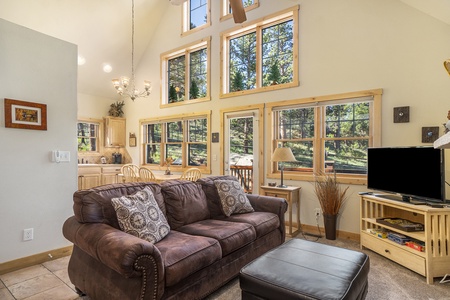 Living room with sofa that seats 3, ottoman, flat screen TV, and windows with view of outdoors.