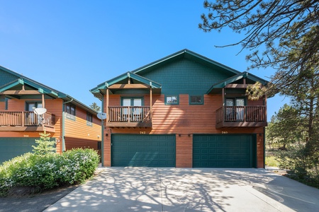 A two-story duplex with wooden siding, featuring two green garage doors and small balconies, surrounded by trees and shrubs.