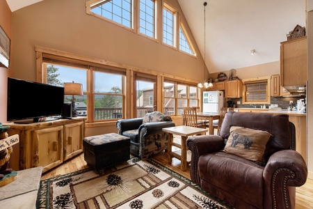 Open living room with 2 arm chairs, one with foot stool, end table, flat screen TV and large windows with view of outdoors.