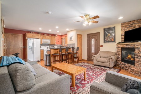 Living room with gray sofas, wooden table, kitchen island with stools, stone fireplace, ceiling fan, and wall art. A dog sits near the kitchen.